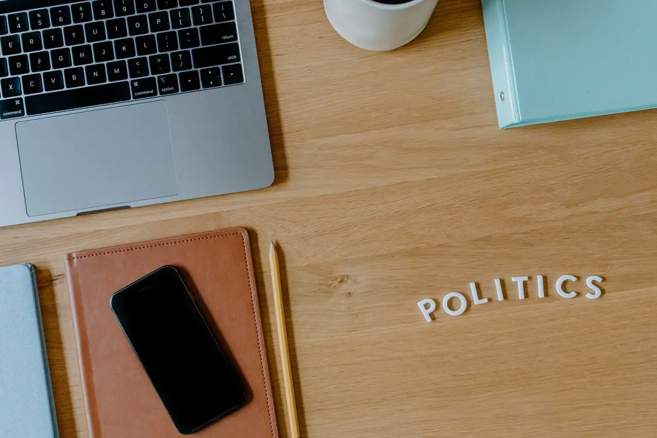 A top view of a workspace featuring a laptop, smartphone, and 'politics' text on a wooden desk.